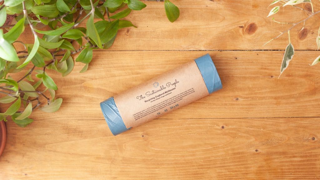 Eine Rolle Zugband-Müllbeutel 35 Liter aus recyceltem Polyethylen von The Sustainable People liegt auf einem Holztisch, in einer Ecke des Bildes sind Blätter zu sehen