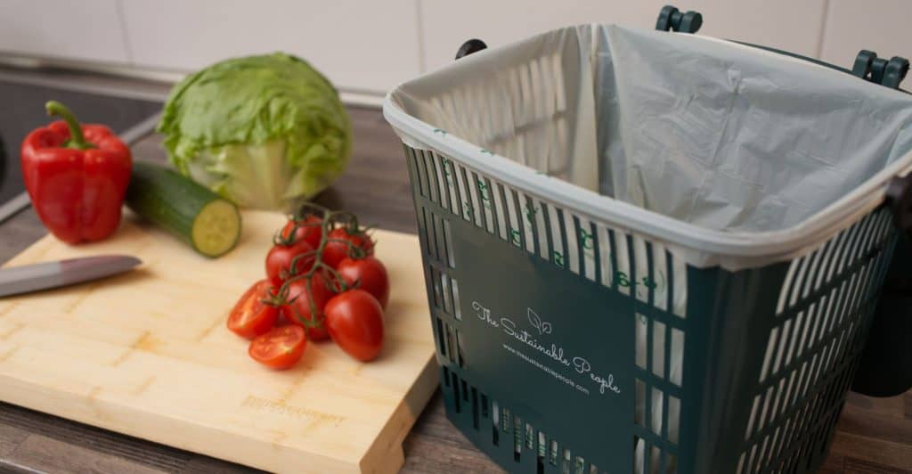 Ein 7,5 Liter Küchen-abfalleimer für Biomüll und Essensreste ausgekleidet mit einem Biomüll-Abfallbeutel steht neben einem Schneidebrett mit Tomaten, einer Gruke und Salat